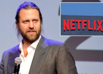 A man with long hair in a suit holding a microphone with a Netflix sign in the background.