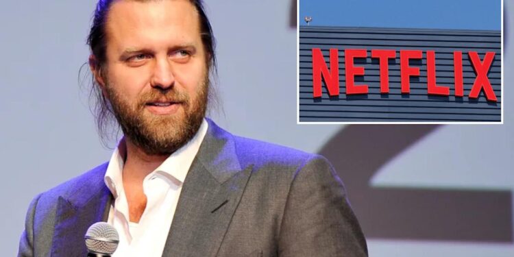 A man with long hair in a suit holding a microphone with a Netflix sign in the background.