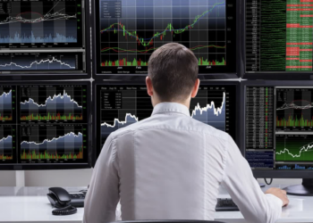 Individual seated at a desk with multiple computer monitors displaying stock market charts and data.