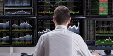 Individual seated at a desk with multiple computer monitors displaying stock market charts and data.