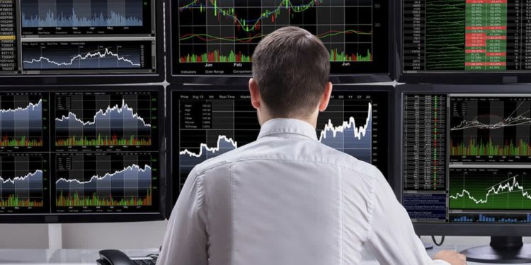Individual seated at a desk with multiple computer monitors displaying stock market charts and data.