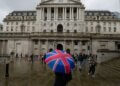 Individual holding a Union Jack umbrella in front of the Bank of England building on a rainy day.