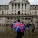 Individual holding a Union Jack umbrella in front of the Bank of England building on a rainy day.