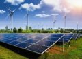 Rows of solar panels in a field with wind turbines in the background under a clear blue sky.