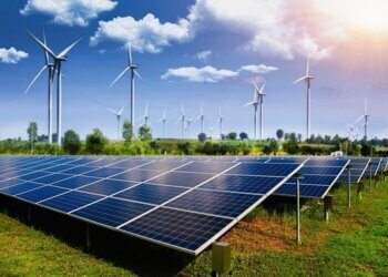 Rows of solar panels in a field with wind turbines in the background under a clear blue sky.