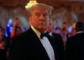 Donald Trump wearing a tuxedo at an event with attendees in the background.