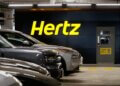 Hertz logo displayed on a wall in a parking garage with parked vehicles in front.