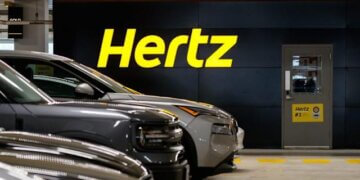 Hertz logo displayed on a wall in a parking garage with parked vehicles in front.