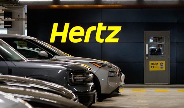 Hertz logo displayed on a wall in a parking garage with parked vehicles in front.