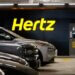 Hertz logo displayed on a wall in a parking garage with parked vehicles in front.