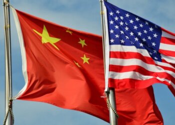 Chinese flag and American flag waving together on flagpoles against a blue sky.