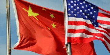 Chinese flag and American flag waving together on flagpoles against a blue sky.