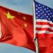 Chinese flag and American flag waving together on flagpoles against a blue sky.