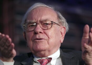 Older man with glasses speaking and gesturing during a public event.