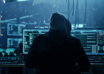 Individual in a hoodie working on multiple computer screens displaying code and data analysis.