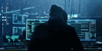 Individual in a hoodie working on multiple computer screens displaying code and data analysis.