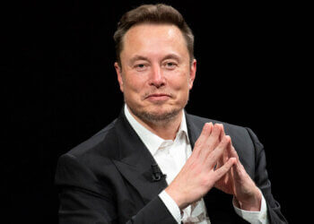 Elon Musk seated with hands clasped, wearing a black suit and white shirt during an interview.