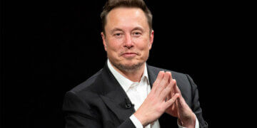 Elon Musk seated with hands clasped, wearing a black suit and white shirt during an interview.