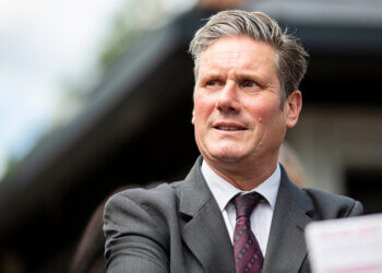 Man wearing a suit with a tie, looking slightly to the side with a neutral expression.