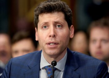 Man in a blue suit speaking into a microphone during a formal hearing setting.