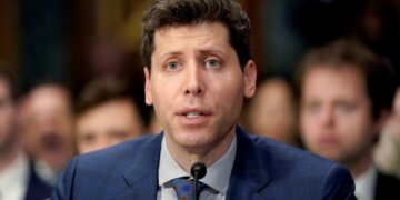 Man in a blue suit speaking into a microphone during a formal hearing setting.