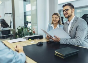 Two professionals in business attire sitting at a table during a meeting with a third person.