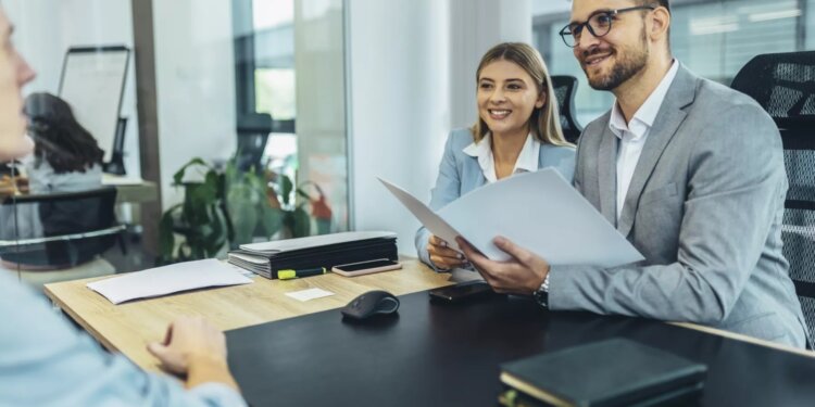 Two professionals in business attire sitting at a table during a meeting with a third person.