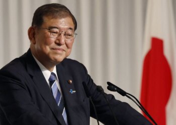 Japanese politician smiling while speaking at a press conference with a flag in the background.