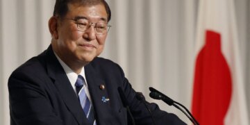 A Japanese politician smiling while standing at a podium during a press conference.
