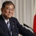 A Japanese politician smiling while standing at a podium during a press conference.