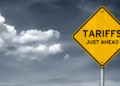 A yellow warning sign with the text 'TARIFFS JUST AHEAD' displayed prominently against a cloudy sky.