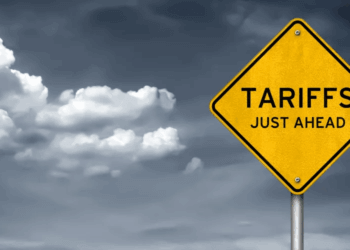 A yellow warning sign with the text 'TARIFFS JUST AHEAD' displayed prominently against a cloudy sky.