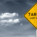 A yellow warning sign with the text 'TARIFFS JUST AHEAD' displayed prominently against a cloudy sky.