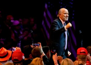 Man in a suit raising his fist while speaking at a public event with an audience in the background.