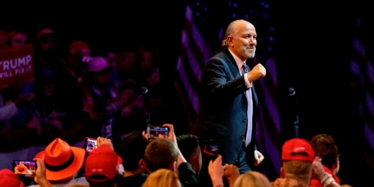 Man in a suit raising his fist while speaking at a public event with an audience in the background.