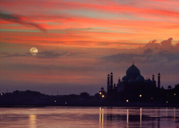 Taj Mahal silhouette against a colorful sunset sky with a full moon visible.