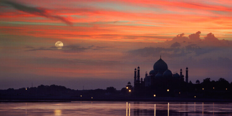 Taj Mahal silhouette against a colorful sunset sky with a full moon visible.