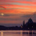 Taj Mahal silhouette against a colorful sunset sky with a full moon visible.
