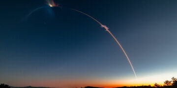 A rocket launch trail creating a bright arc in the evening sky against a gradient backdrop.