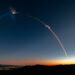 A rocket launch trail creating a bright arc in the evening sky against a gradient backdrop.