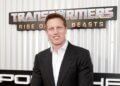 Man in a black suit standing in front of a Transformers movie premiere backdrop.