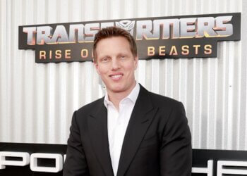 Man in a black suit standing in front of a Transformers movie premiere backdrop.
