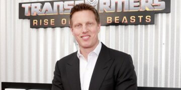 Man in a black suit standing in front of a Transformers movie premiere backdrop.