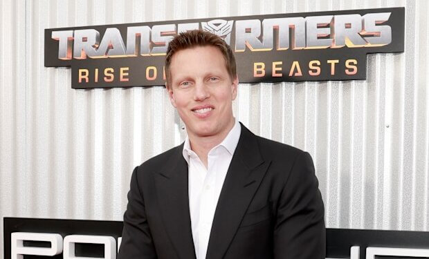 Man in a black suit standing in front of a Transformers movie premiere backdrop.
