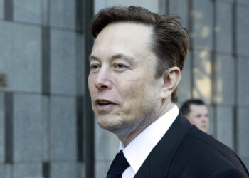 Man with short brown hair wearing a black suit and tie, standing outdoors with a neutral expression.