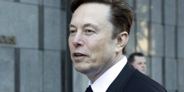 Man with short brown hair wearing a black suit and tie, standing outdoors with a neutral expression.