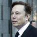 Man with short brown hair wearing a black suit and tie, standing outdoors with a neutral expression.
