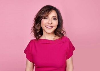 Woman with shoulder-length brown hair wearing a pink top against a pink background.
