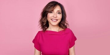 Woman with shoulder-length brown hair wearing a pink top against a pink background.