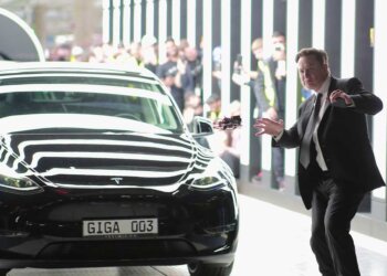 Elon Musk gesturing near a black Tesla vehicle in a modern setting.